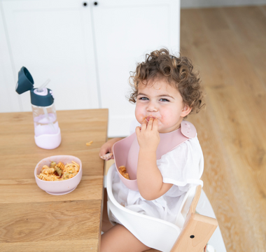 Toddler eating pasta with pink mess-free bib at wooden table, food-safe catch-all design