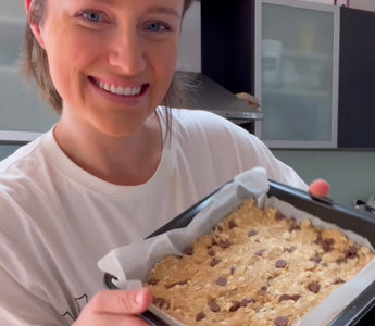 Woman smiling while holding a baking tray with chocolate chip oat bars, ready to bake.