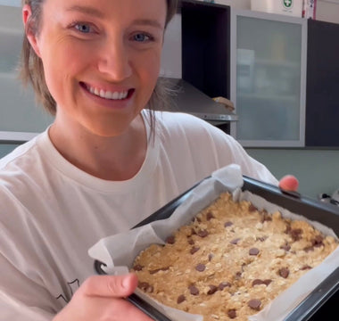 Woman smiling while holding a baking tray with chocolate chip oat bars, ready to bake.